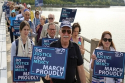 People marching over a bridge holding signs that read DO JUSTICE LOVE MERCY MARCH PROUDLY
