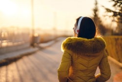 Women wearing a yellow coat facing away from the camera wearing a pair of headphones and looking out over a dock