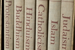 Books on a shelf with different titles on the spines: Judaism, Islam, Catholicism, Hinduism, Buddism, Protestantism