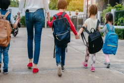 Seen from behind: four young children with backpacks walking hand-in-hand with each other and a mother or caregiver