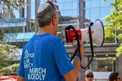 A man facing away from the camera holding a megaphone and wearing a shirt that reads DO JUSTICE LOVE MERCY WALK PROUDLY