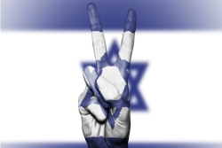 Hand giving a peace sign and painted to match the Israeli flag which is waving in the background