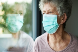 Older woman staring out a window while wearing a face mask