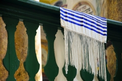 Blue and white Jewish prayer shawl laying on a pew with Hebrew visible on the wall in the background