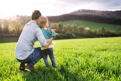 Father squatting to hold toddler in open grassy expanse with mountains in the background