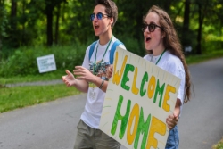 Two enthusiastic camp counselors holding a hand painted sign that reads WELCOME HOME