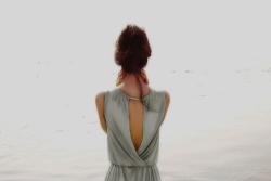 A brunette woman from the back with her hair up and her hands on her neck as she looks out over a body of water