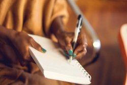 Hand of a Black woman wearing blue nail polish and using a pen to write in a notebook as if penning a letter