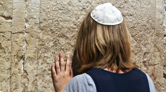 Woman wearing a kippah turned away from the camera with her hand and forehead resting against the Western Wall 